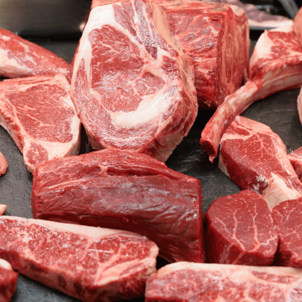 A selection of raw beef cuts displayed on a black surface, featuring various types including ribeye steaks and chunks of brisket. The marbling and fresh red color of the meat are clearly visible.