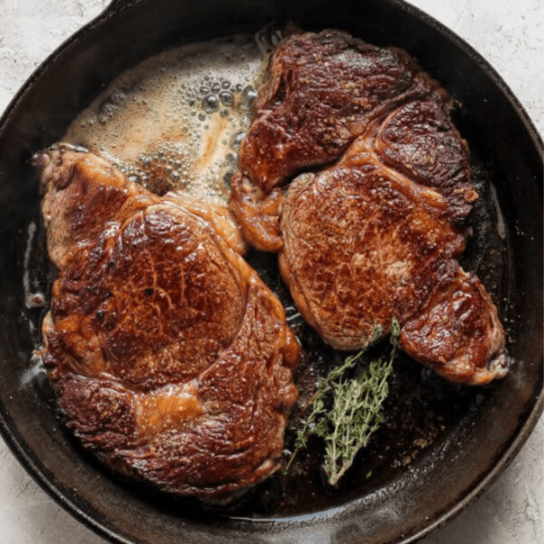 Two thick, juicy steaks with a rich brown crust are sizzling in a cast iron skillet. Juices and melted butter pool around the steaks, and a sprig of fresh thyme is nestled alongside, adding a hint of green to the scene.