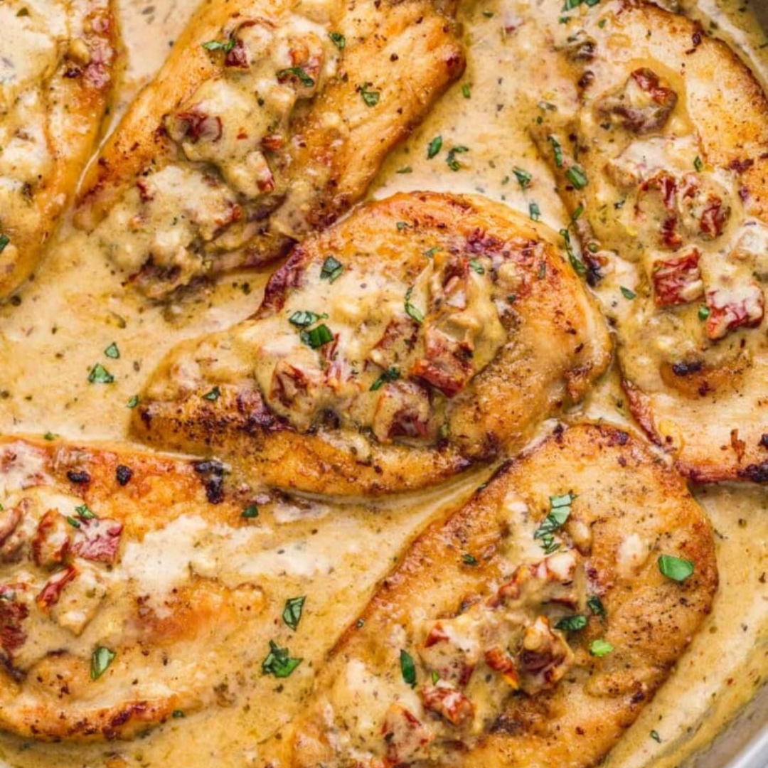 A close-up of a skillet filled with creamy Tuscan chicken. The dish features golden-brown chicken breasts smothered in a rich, creamy sauce with chunks of sun-dried tomatoes, garlic, and fresh herbs. The sauce appears to be infused with a blend of spices.