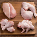 A wooden cutting board displaying four cuts of raw chicken arranged separately: a skinless chicken breast on the top left, a chicken leg quarter on the top right, two drumsticks on the bottom left, and a whole chicken wing on the bottom right.