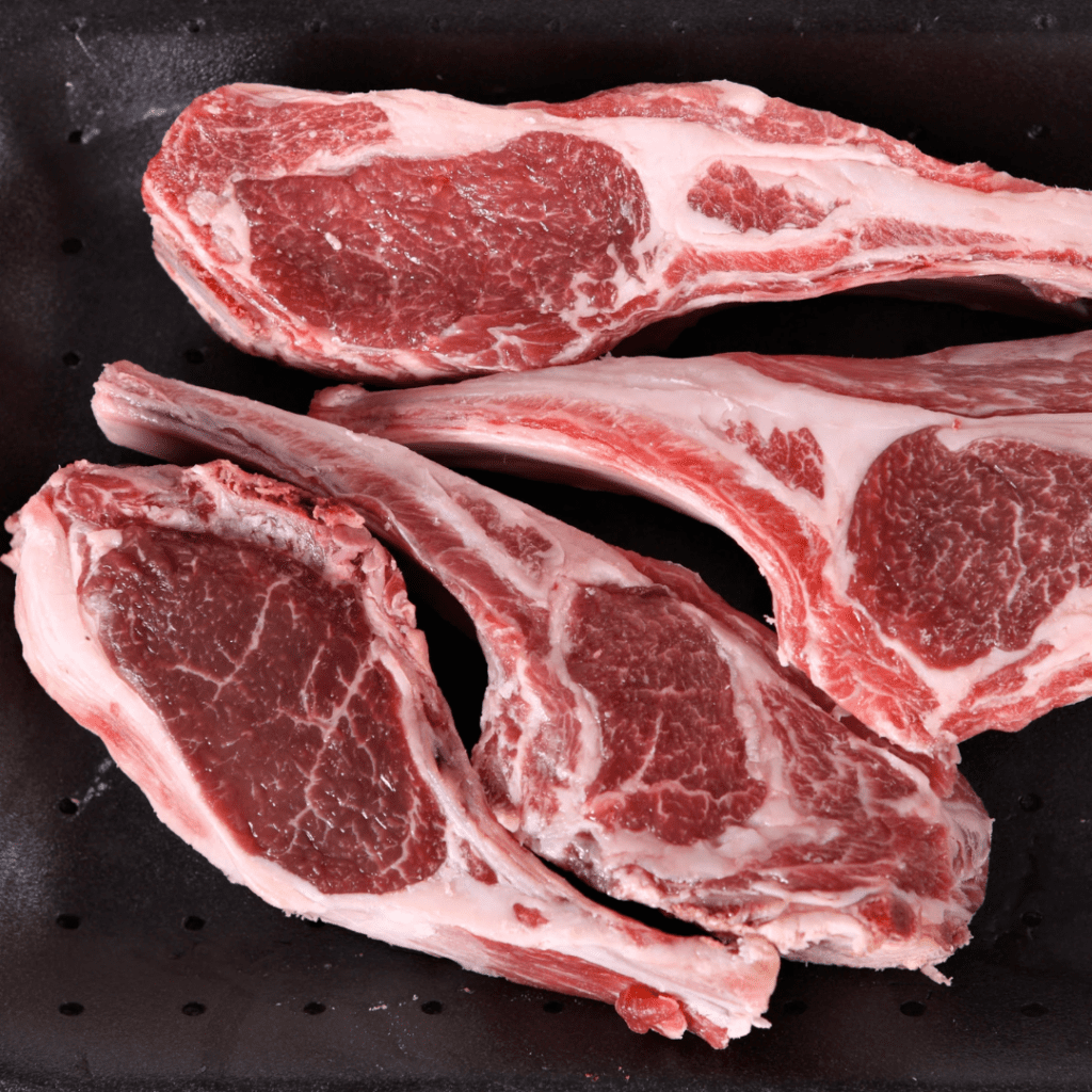 Raw lamb chops with marbled fat are arranged closely together on a black surface. The red meat and white fat create a visually appealing contrast. The lamb chops have a fresh and uncooked appearance.