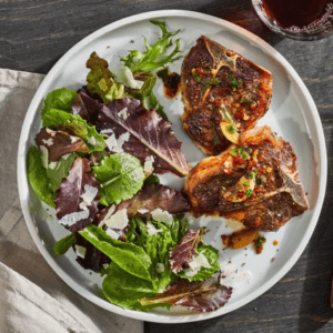 A white plate features two seasoned and garnished lamb chops alongside a fresh green salad topped with shaved Parmesan cheese. A glass of red wine and a beige napkin are partially visible next to the plate. The background has a dark wood texture.