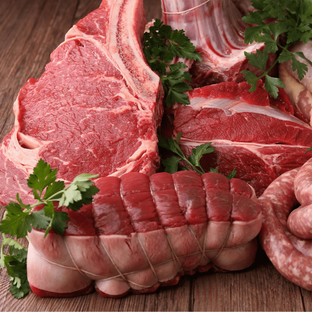 A display of various cuts of raw red meat, including a T-bone steak, ribeye, roast, and sausages, arranged on a wooden surface. Fresh parsley is garnished throughout to enhance presentation.