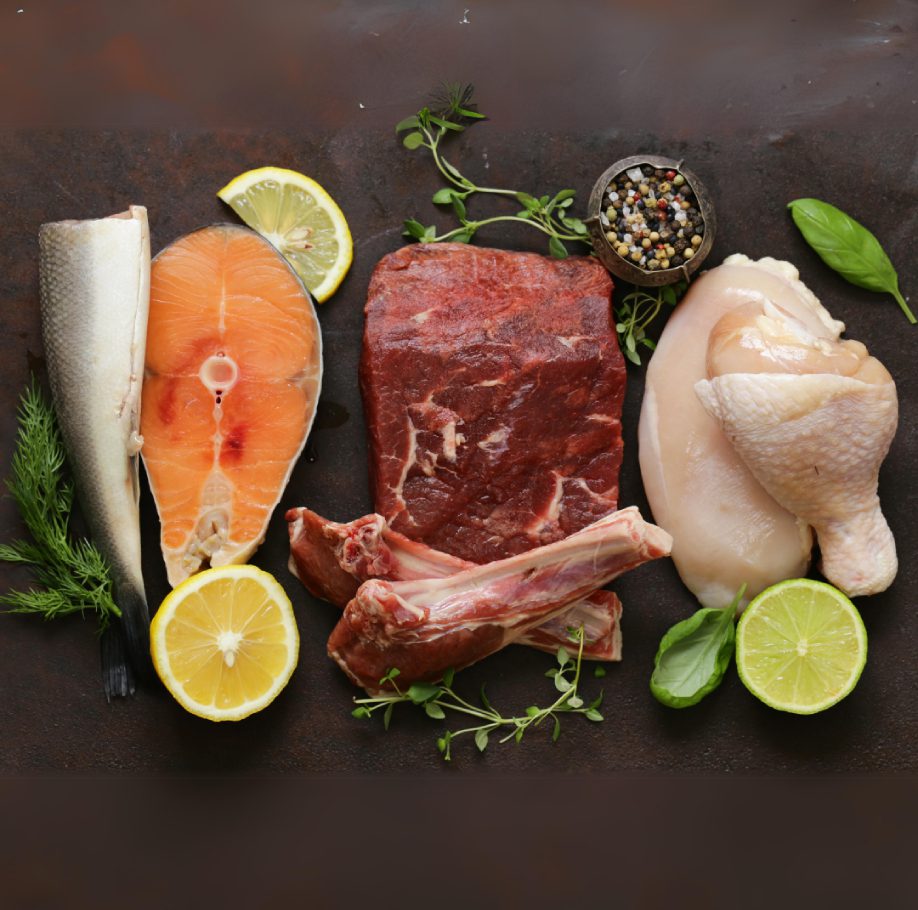 A selection of raw meat and fish: a salmon steak with a lemon slice, a fish filet with dill and lemon, a beef steak, lamb chops, and chicken breast with basil and lime. A small bowl of mixed peppercorns and fresh herbs are arranged around the meats.