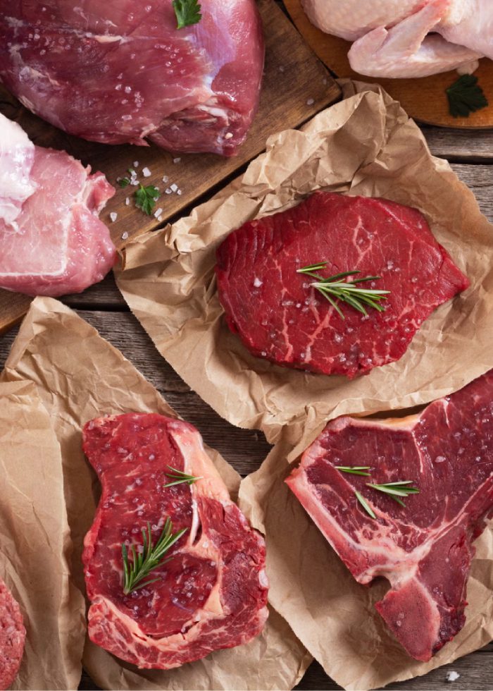 Various cuts of raw meat are displayed on parchment paper. The meats include steaks garnished with rosemary, a piece of pork, and chicken. The scene is set on a rustic wooden surface.