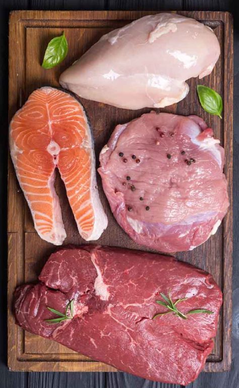 A wooden cutting board displays four types of raw meat and fish: a chicken breast, a salmon steak, a piece of pork sprinkled with black peppercorns, and a beef steak garnished with rosemary. Small green leaves are scattered around the meats.