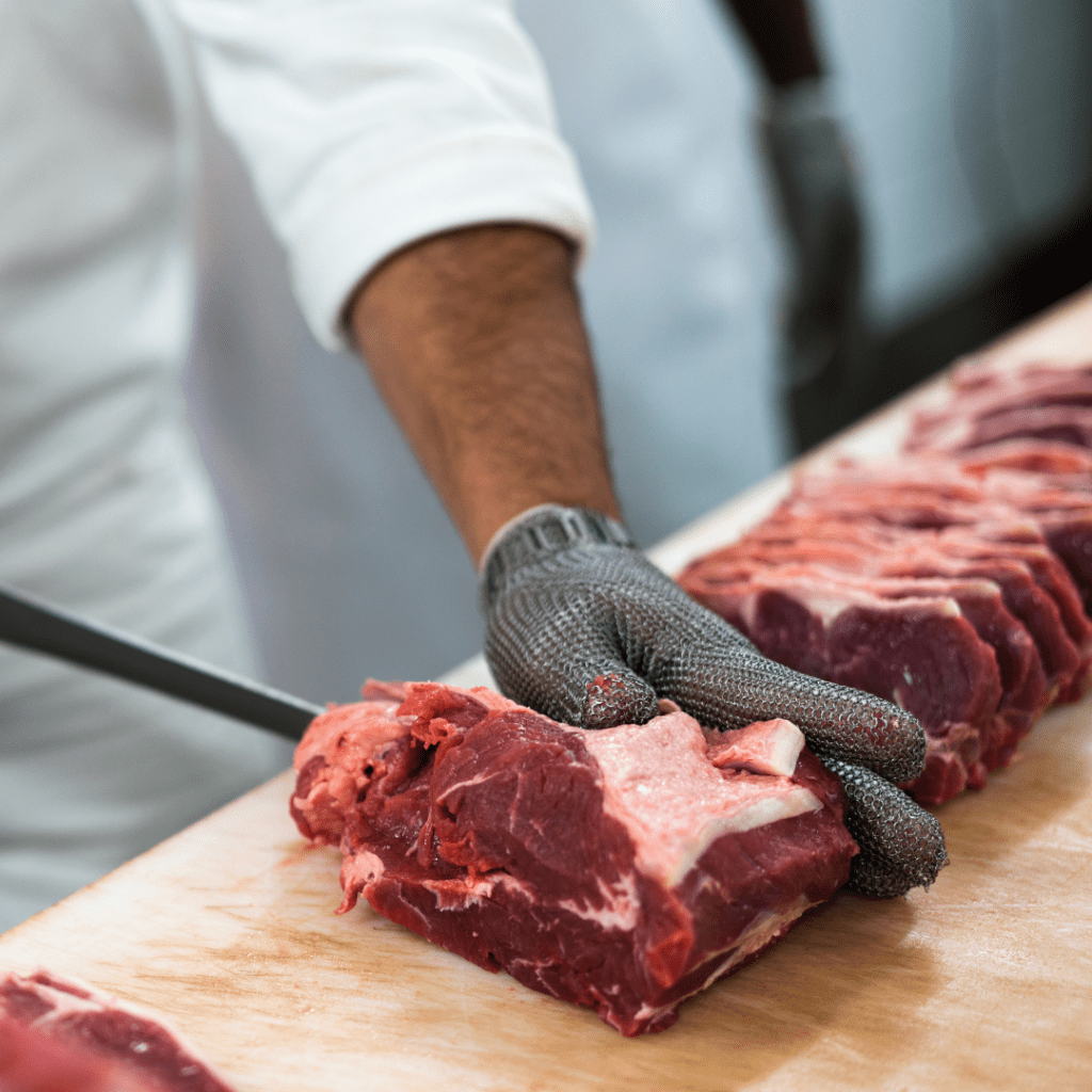 A person wearing a white uniform and a cut-resistant glove is slicing raw meat with a knife on a wooden cutting board. Several slices of red meat are arranged in a row on the board.