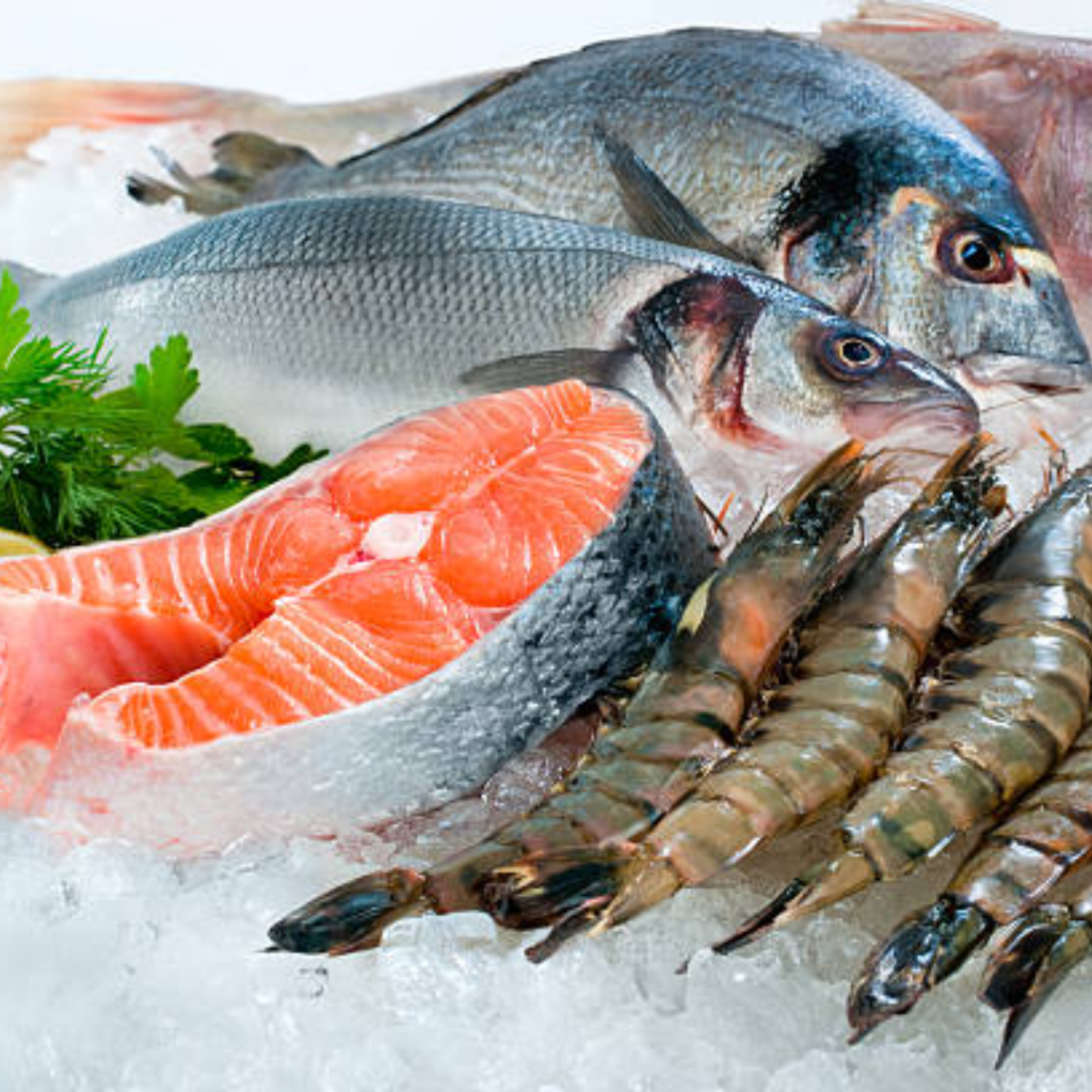 A selection of fresh seafood on ice, including raw salmon fillets, whole fish, and large prawns. There is a garnish of fresh parsley on the ice, adding a touch of greenery to the display. The seafood appears fresh and ready for cooking.