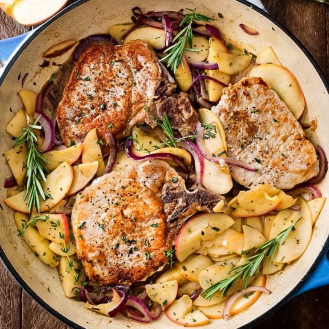 A skillet containing two browned pork chops, surrounded by slices of apples and red onions, all garnished with sprigs of rosemary and a sprinkle of chopped parsley.