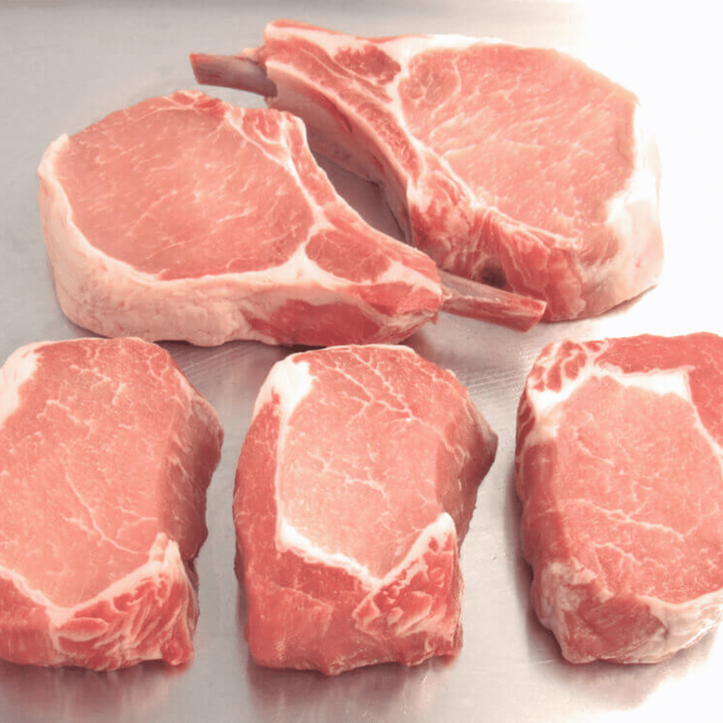 Several cuts of raw pork chops are displayed on a metallic surface. The cuts vary slightly in shape and size, with some showing bone-in sections. The meat has visible marbling and a light pink color, indicating its uncooked state.