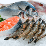 A display of fresh seafood on a bed of ice, including whole fish, salmon fillets, and large prawns. There's a sprig of parsley on the left for garnish. The seafood is vibrant and glistening, indicating freshness.