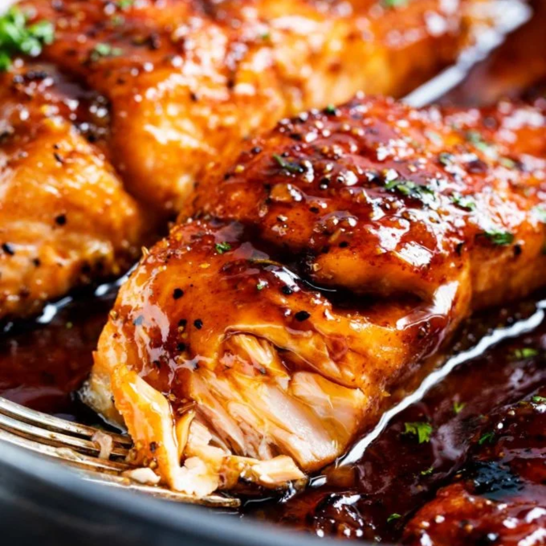 Close-up of glazed salmon fillets garnished with herbs in a dark, rich sauce. One fillet is being flaked with a silver fork, revealing the tender, flaky interior. The glaze is glossy and thick, enhancing the vibrant orange color of the salmon.