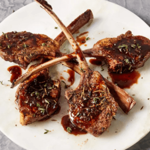 A white plate with four grilled lamb chops garnished with fresh herbs and drizzled with a dark, rich sauce. The lamb chops have a crispy, well-seasoned crust and are presented elegantly.