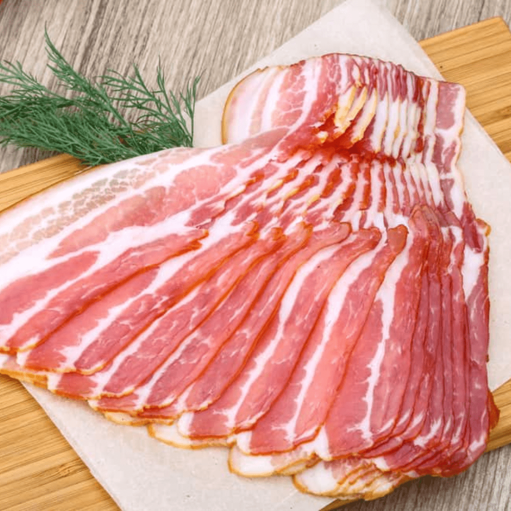 A stack of uncooked, thinly sliced bacon is neatly arranged on a cutting board. The pink and white marbling of the bacon is prominent, and a sprig of fresh dill lies next to the bacon slices. The background features a wooden table surface.