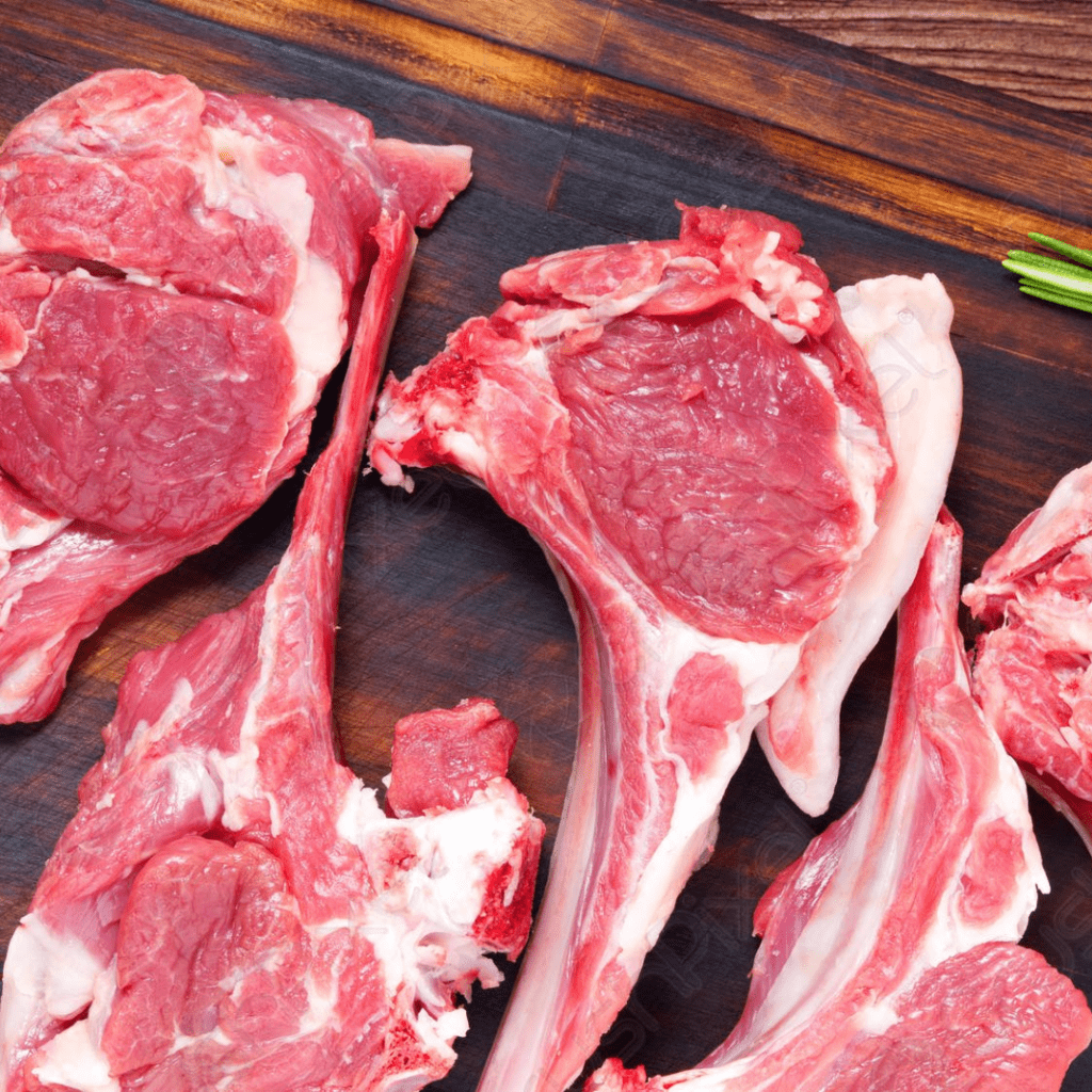 A close-up photograph of raw lamb chops displayed on a dark wooden cutting board. The meat is pink and marbled with white fat, with several chops placed at various angles. A few sprigs of fresh herbs can be seen on the side of the board.