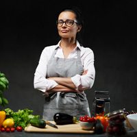 A woman wearing glasses and a gray apron stands confidently with her arms crossed in a kitchen. Fresh vegetables including an eggplant, zucchini, tomatoes, lettuce, and bell peppers are laid out on a cutting board and counter in front of her.